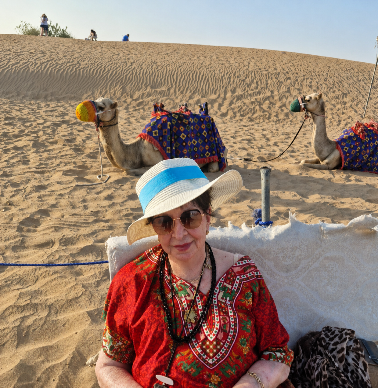 Marlene Galeazzi sentada no deserto de Dubai com camelos ao fundo.